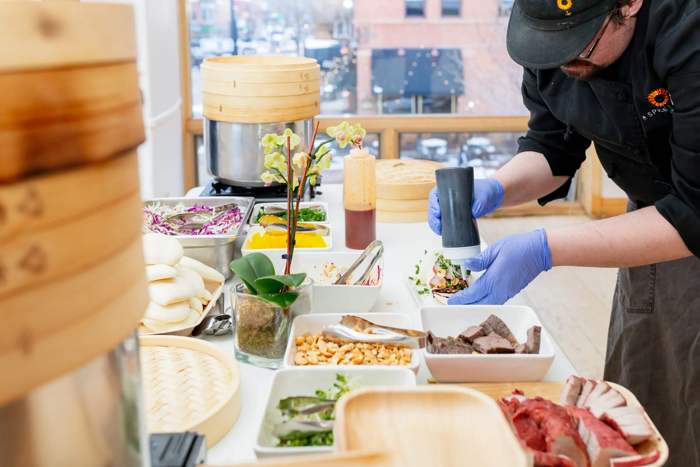 Two A Spice of Life chefs in gloves building bao buns at a live station in a downtown window, with the street behind them