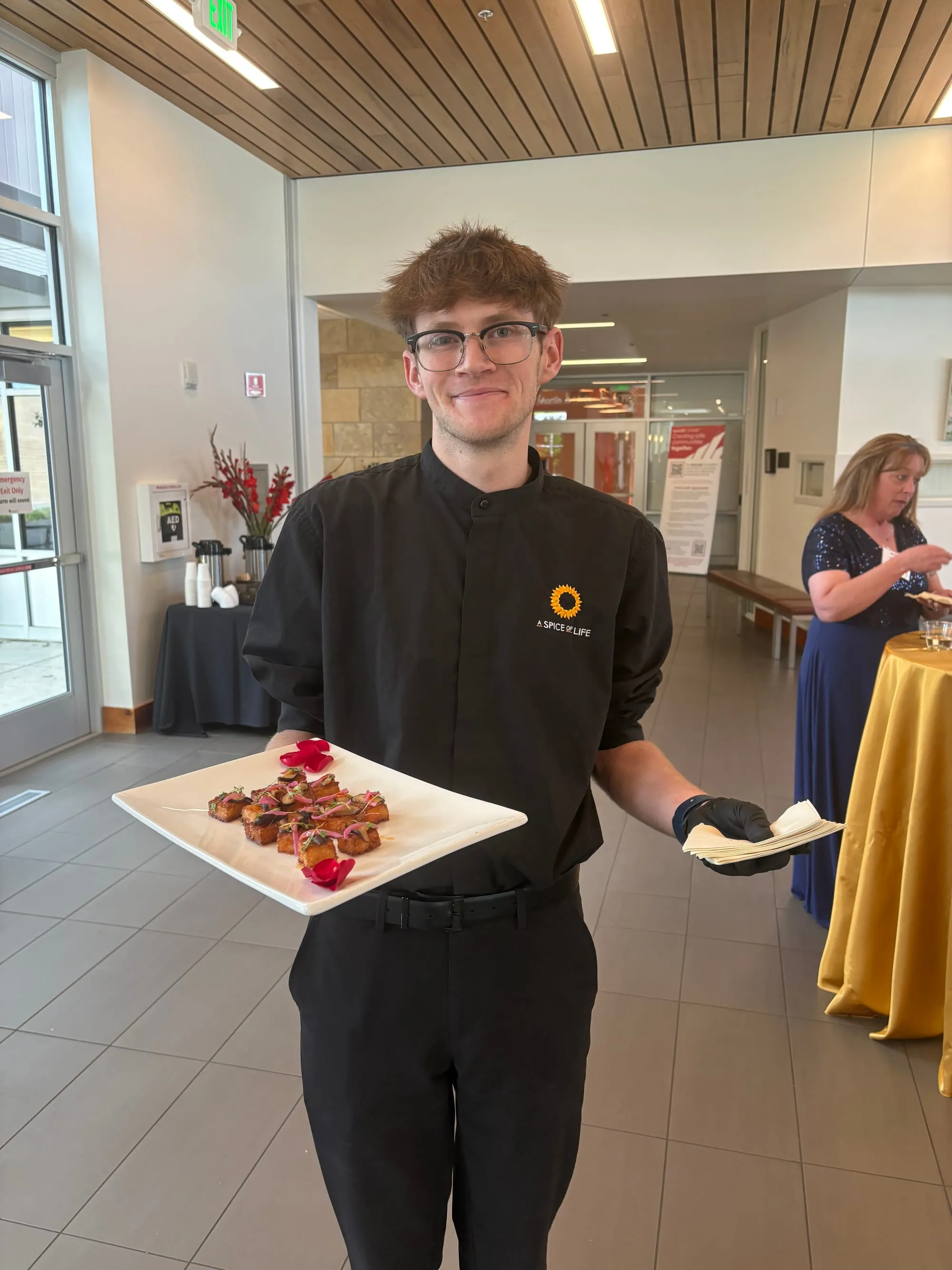 An A Spice of Life server in uniform presenting a tray of plated appetizers in a bright venue lobby